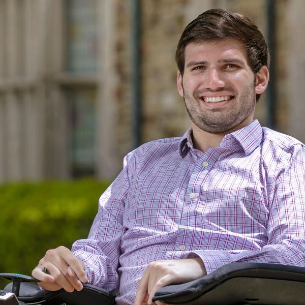 A dark haired young man with a wide smile looks into the camera from his motorized wheelchair. 