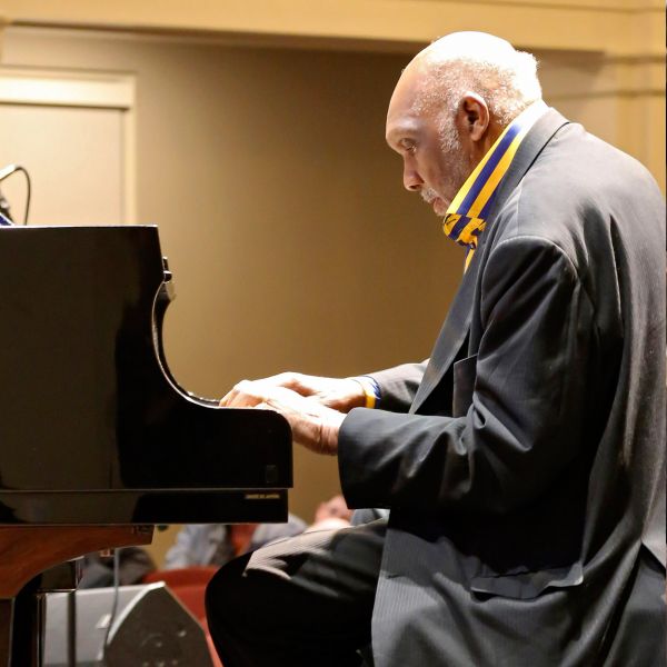 A white haired man sits in profile at a grand piano with his hands resting on the keys.