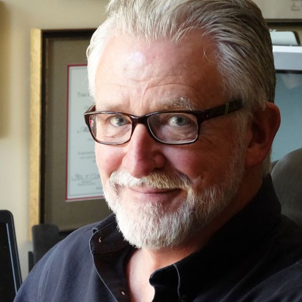 A man with a goatee and square glasses frames sits in dramatic window light.