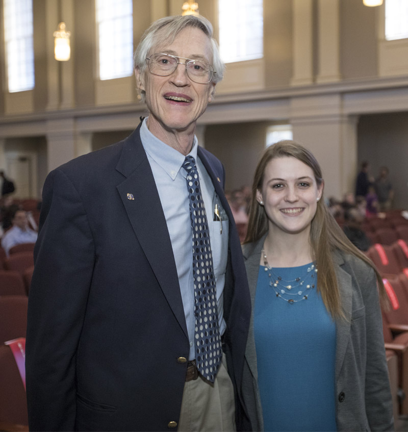 A young woman in blue stands next to a tall gentleman in a sports coat and tie. Both smile into the camera.