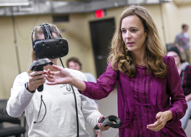A woman in a maroon top guides the hand of a student who is wearing a VR headset.