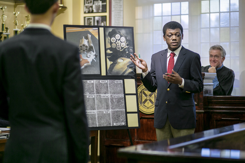 An older man, Professor Pohlmann, sits on the bench as a student in a suit points at a poster of evidence