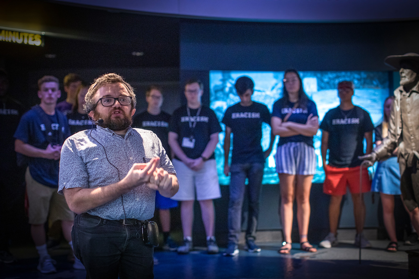 Bearded man in foreground speaks as students in background listen in museum