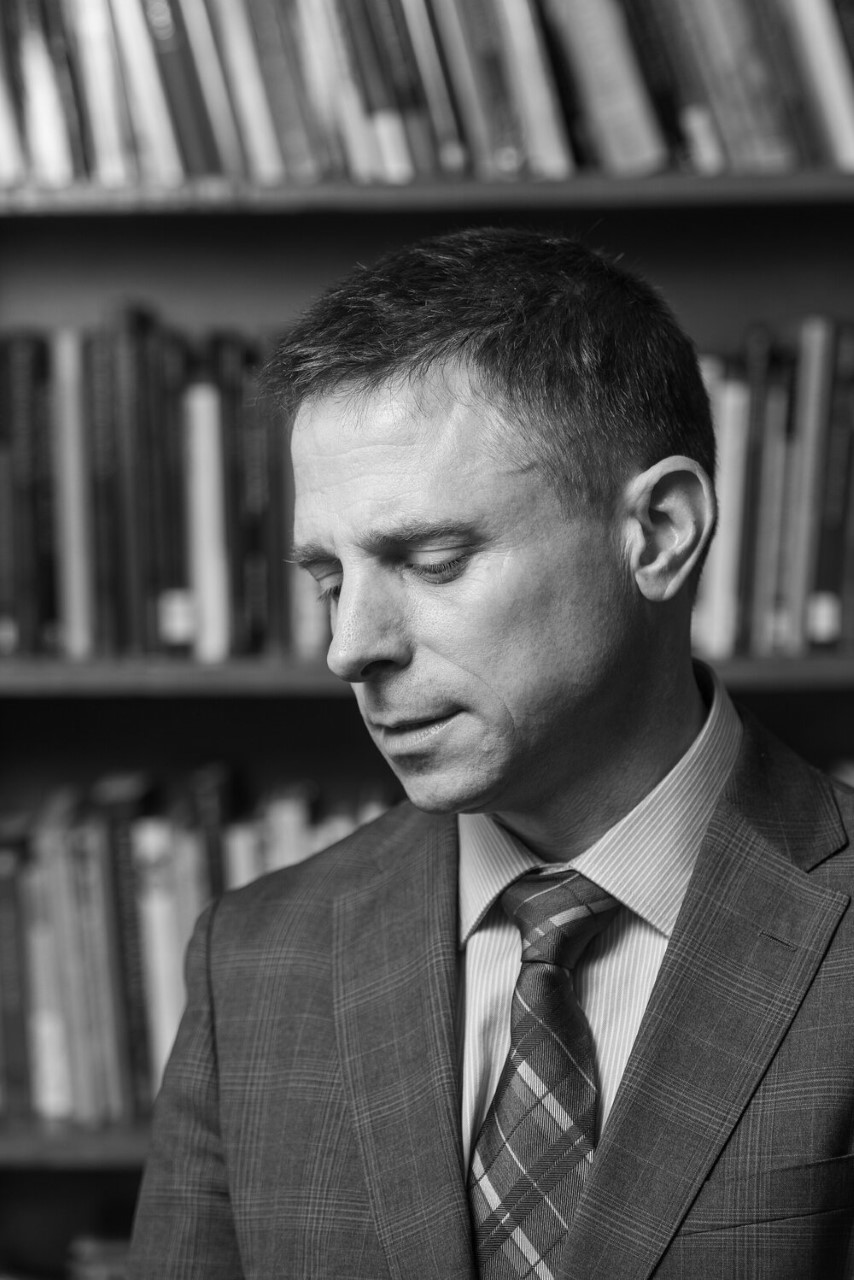 A black and white photo of a professor in suit and tie standing in is library.