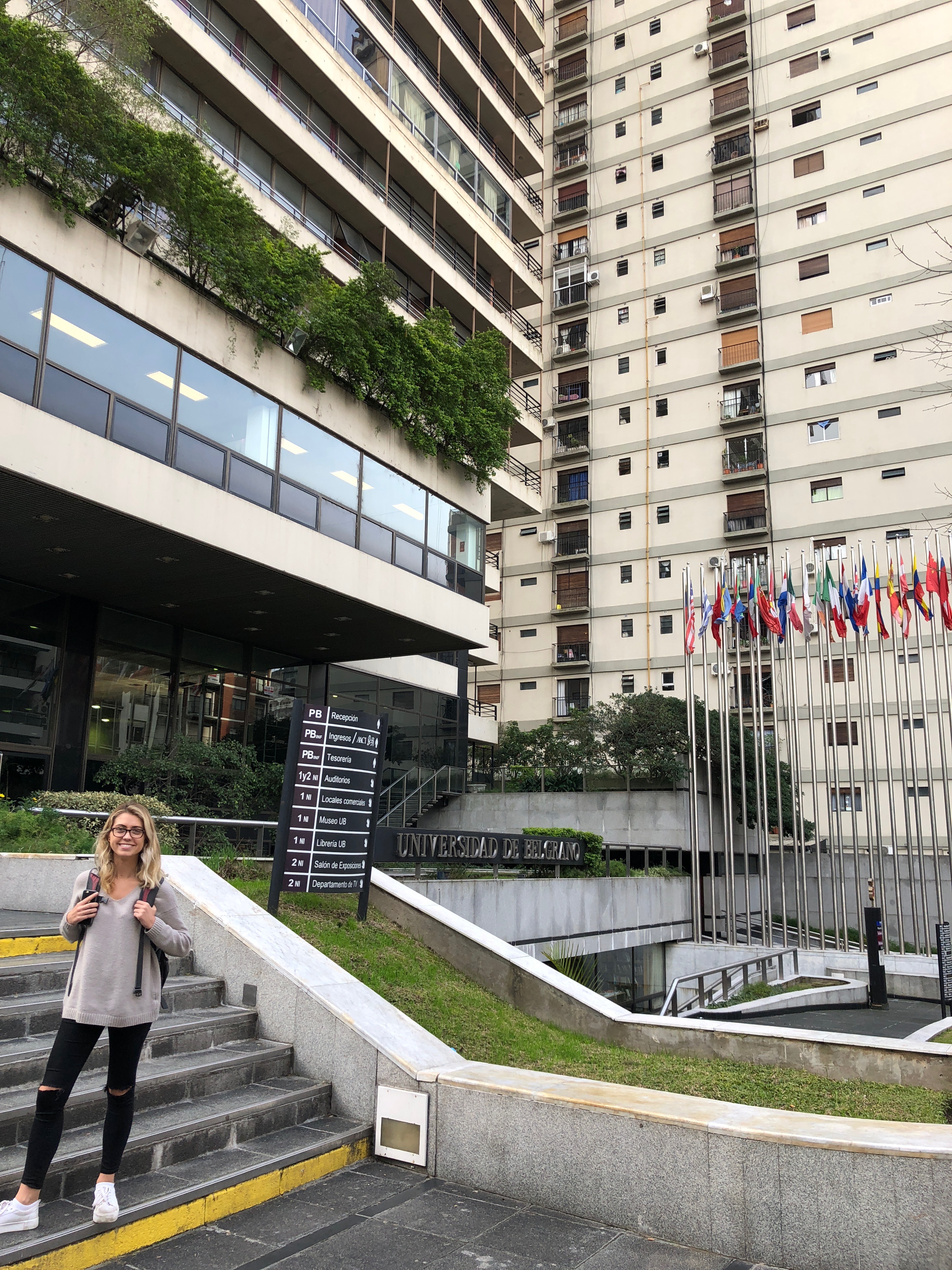 a yoiung woman with a backpack stands in front of a building with many flags 