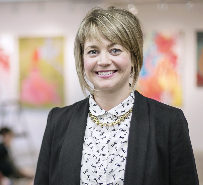 A woman with short blonde hair smiles in front of a collection of paintings.