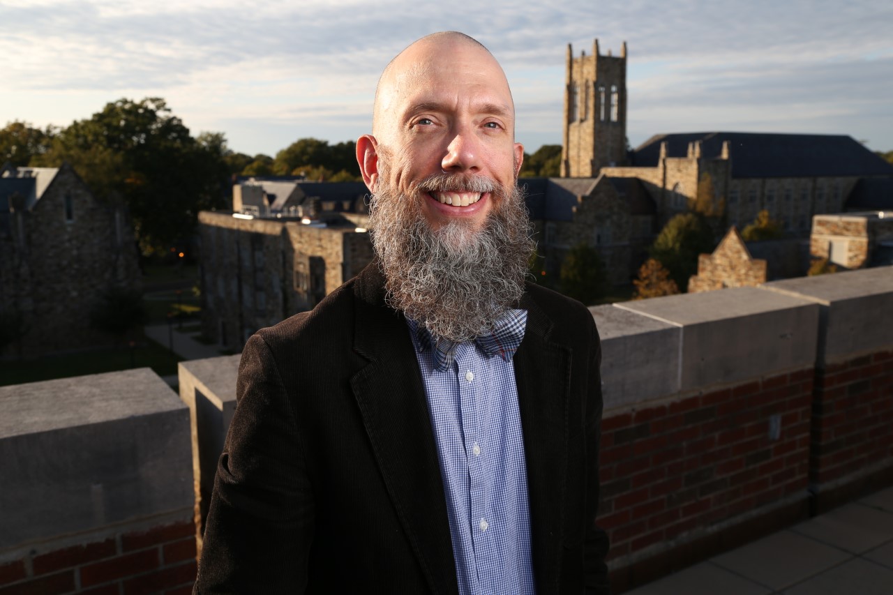 A bearded professor with a bowtie stands atop a building.