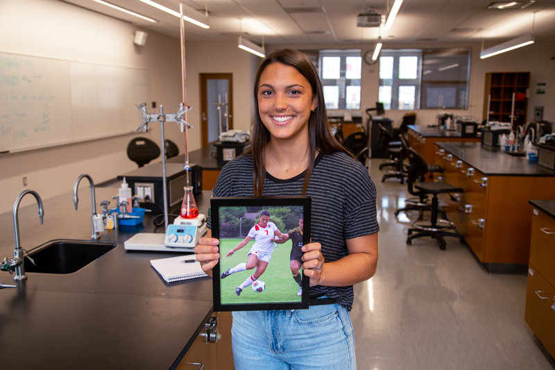a young woman in a lab holds a photo of herself playing soccer