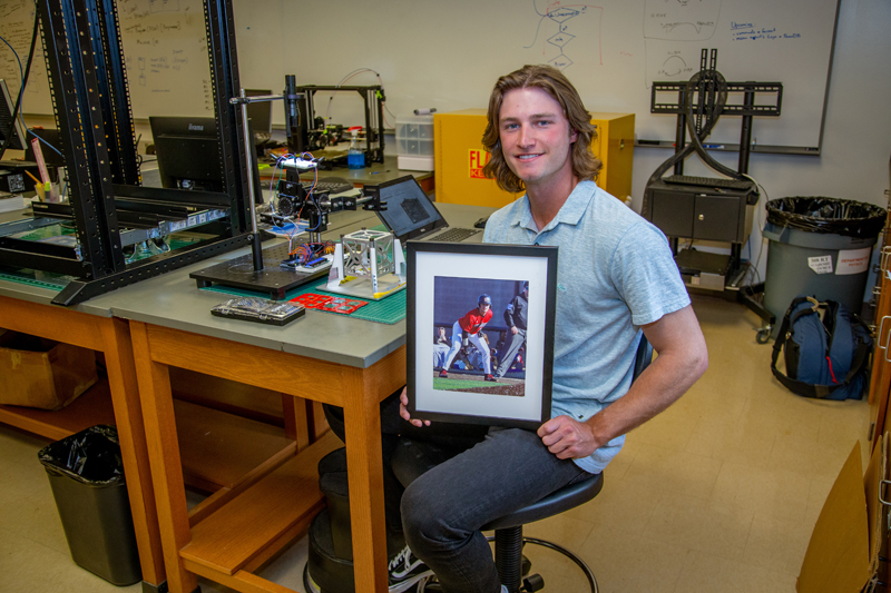 a young man in a physics lab hold a photo of himself playing baseball