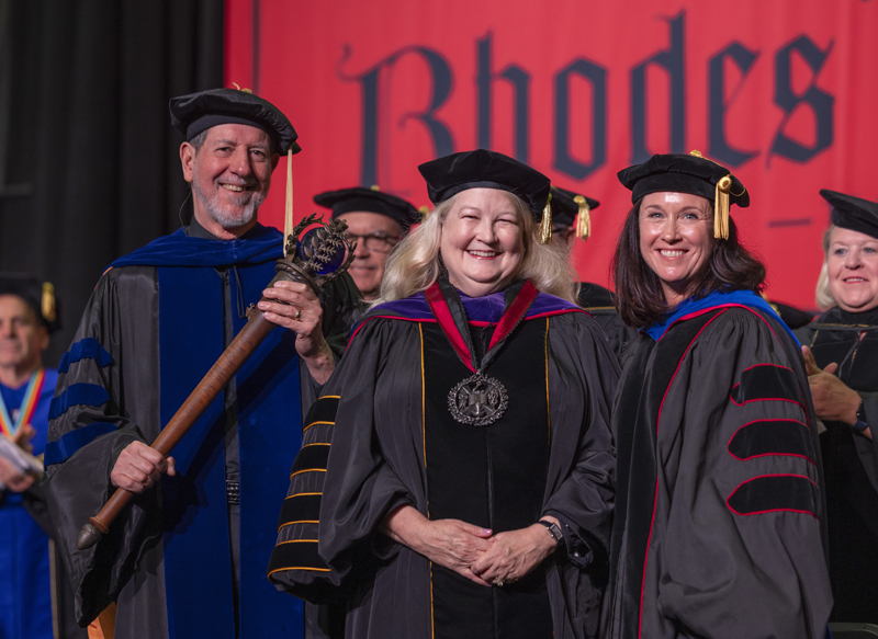 a woman in academic robes flanked by a male and female, both in academic robs