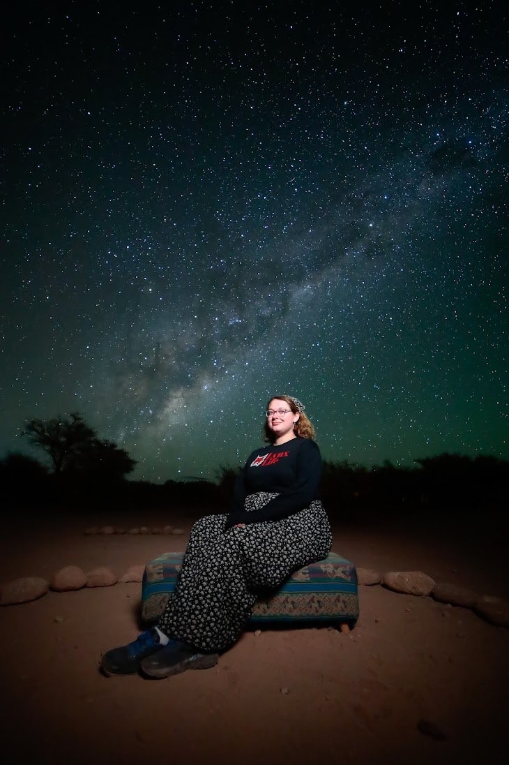 a young woman sits in a desert stone circle with a brilliant starry sky behind her
