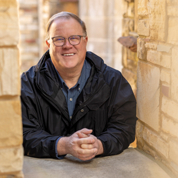 a man with glasses leans on a stone sill