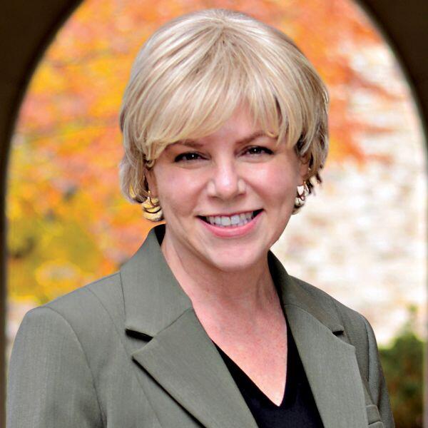 A woman smiles into the camera with beautiful fall foliage behind her.