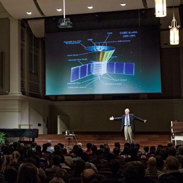 A man in suit and tie stands on stage in front of a crowded room with a space probe projected on a screen above him.