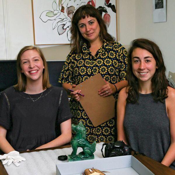 Two students seated at a table strewn with ancient artifacts with an instructor standing behind them.