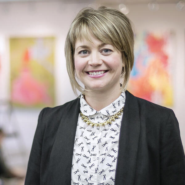 A woman with short blonde hair smiles in front of a collection of paintings.