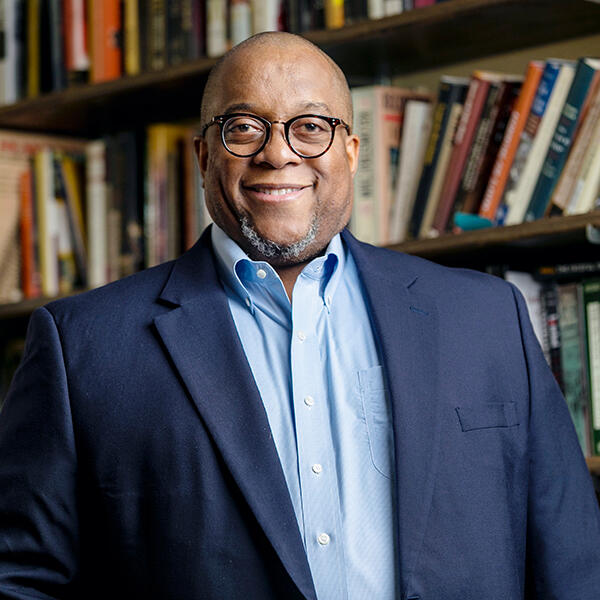 an African American man in a blue suit stands before a bookcase