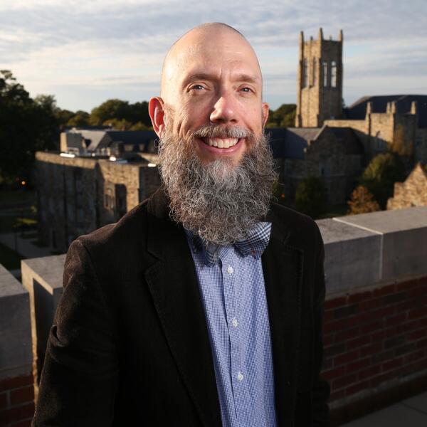 A bearded professor with a bowtie stands atop a building.