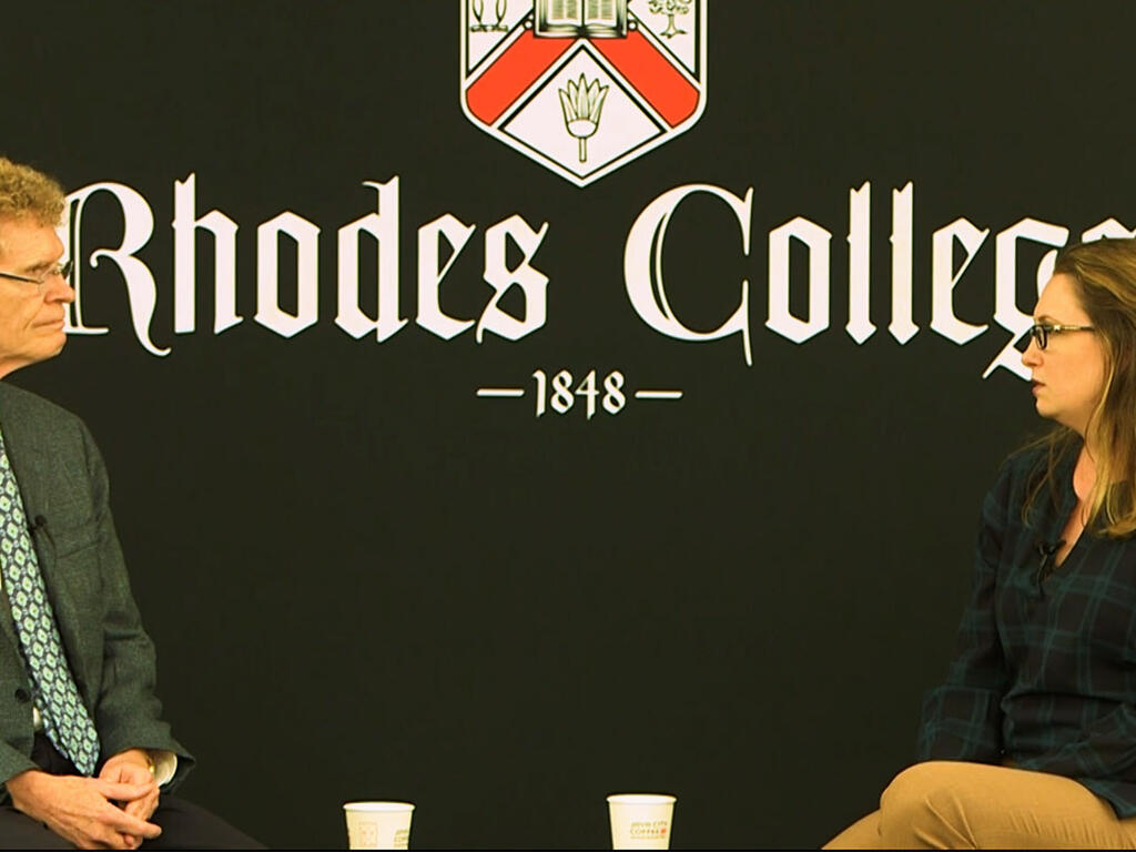 A man and woman sit across from each other in front of a black backdrop that displays the Rhodes College logo.