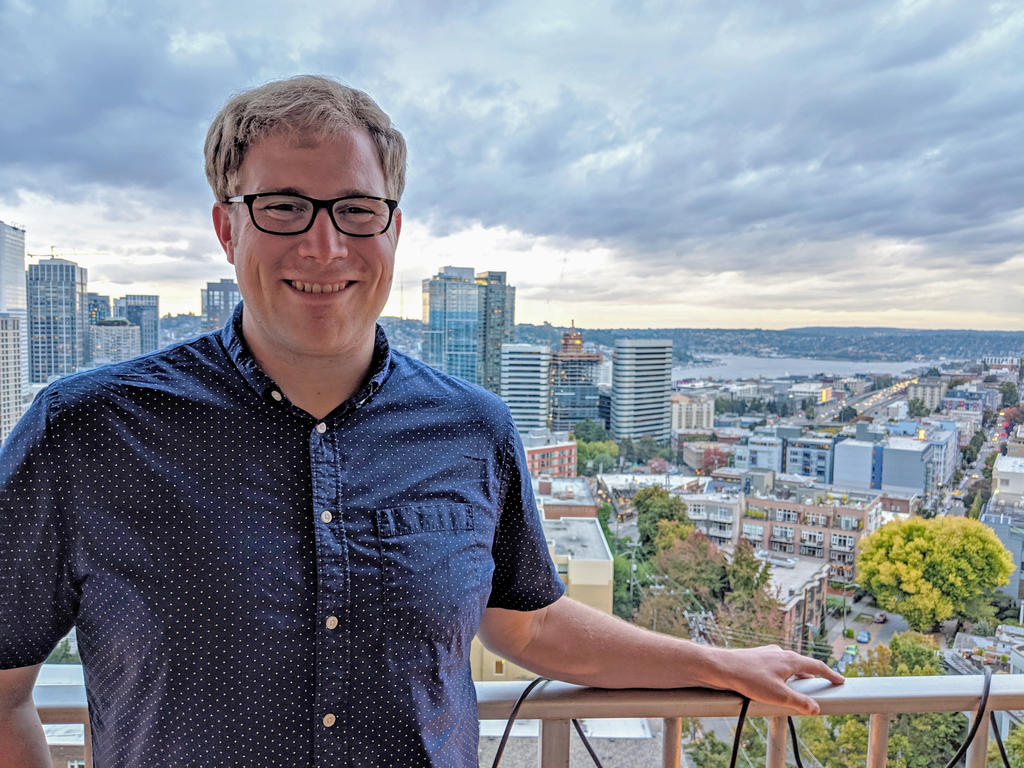 Man looks at camera on balcony in front of a partial Seattle skyline on a cloudy day.