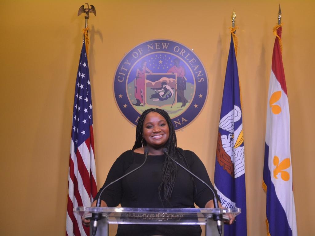 A young woman stands in front of a podium with flags and a seal in the background