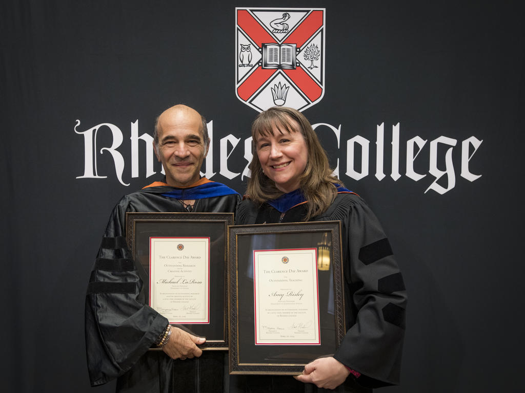 two college professors holding awards standing in front of black backdrop with a college logo printed on it