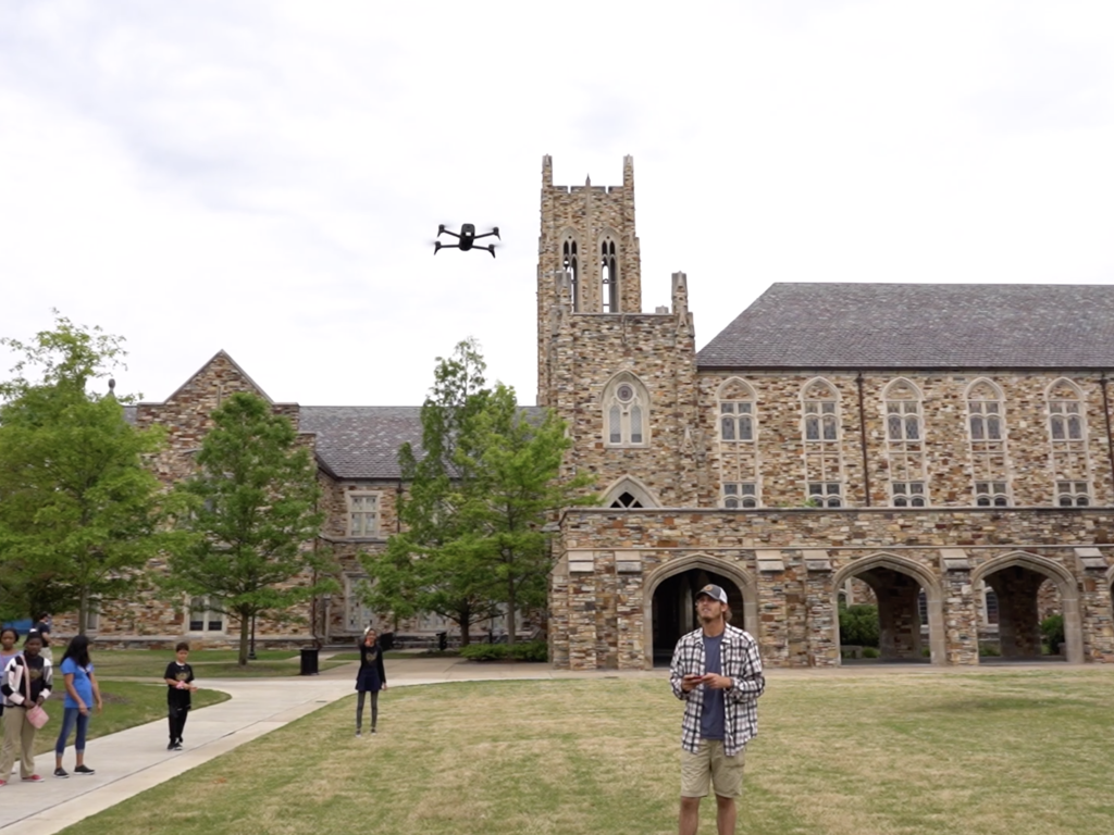 A student flies a drone in front of an audience, flanked by a stone building 