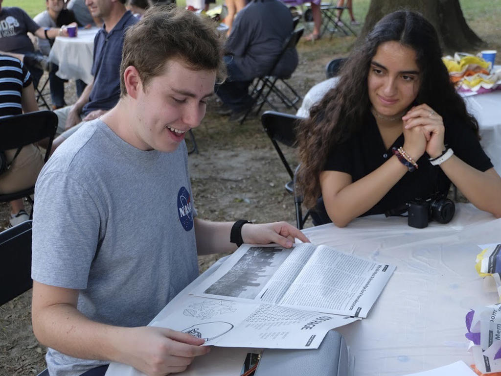 a young man seated at a table, holding a newspaper as a young woman looks on