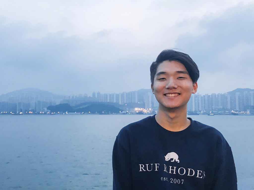 a young Asian male stands in front of a harbor surrounded by hills