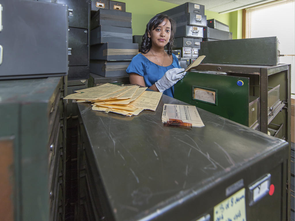 a young woman with gloves on and surrounded by file cabinets looks at the camers