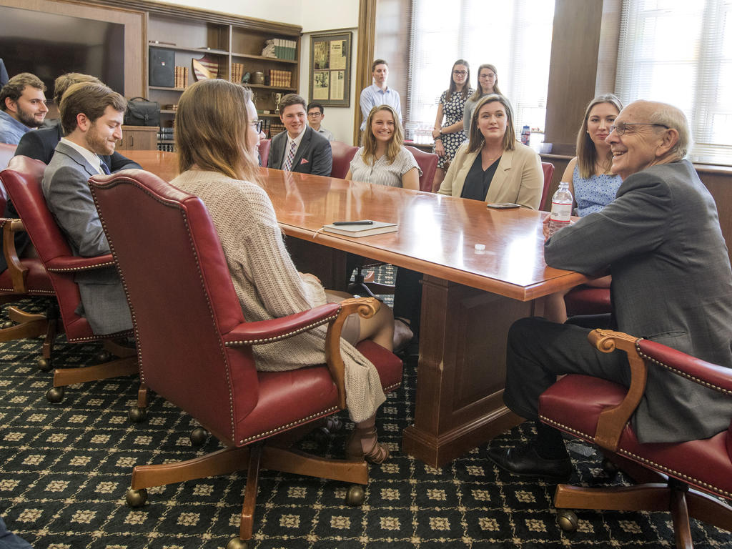 an older man seated at a table leans forward in conversation with a group of students