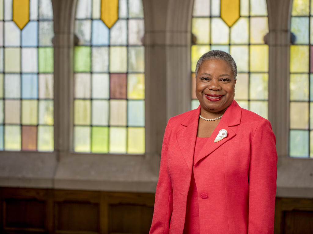 A person in a red suit smiles at the camera in front of stained glass windows