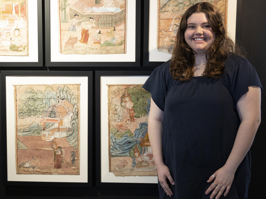 a young woman stands in front of Thai paintings