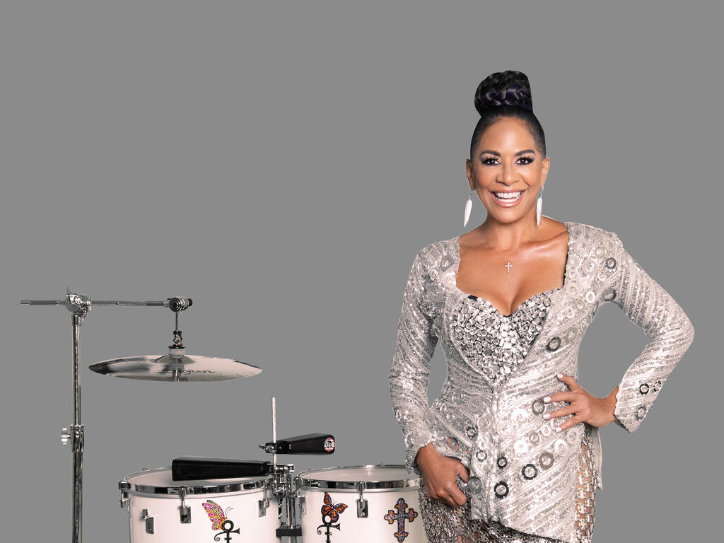 A woman in a silver dress poses next to a partial drum kit.