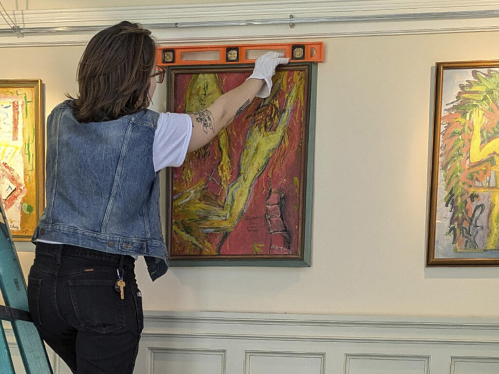 a woman on a ladder hangs artwork in a gallery