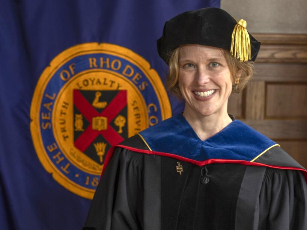 head and shoulder image of Caki Wilkinson in academic cap and gown, standing in front of Rhodes College banner