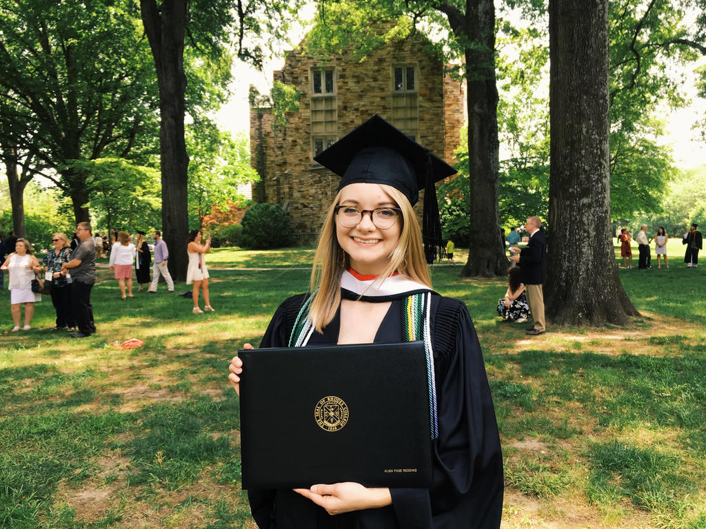 college student in cap in gown standing among trees, holding diploma