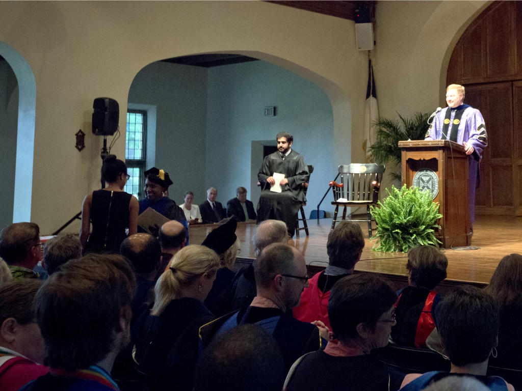 an awards ceremony taking place, with a professor behind the podium and a student anxiously sitting in the adjacent chair