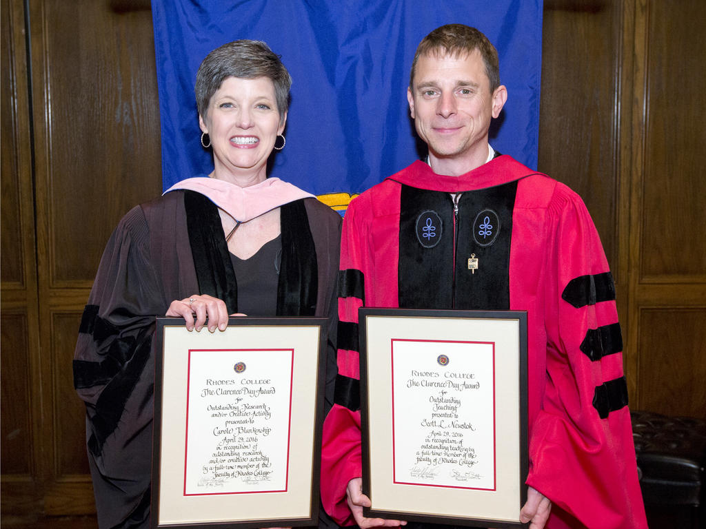 two professors in academic robes holding their awards proudly in front of a blue backdrop