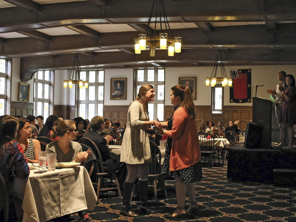 students seated in the cafeteria; one student is receiving an award while her peers clap for her