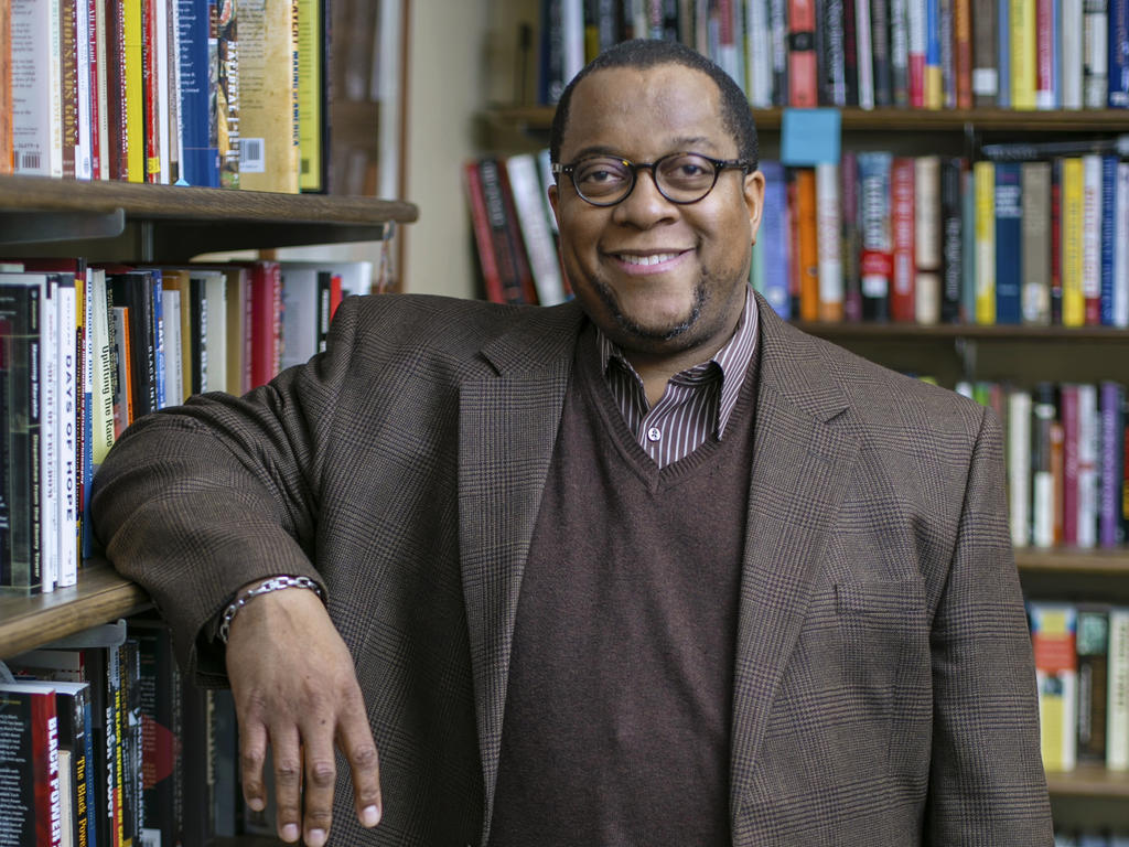 an African American male professor leaning against a bookshelf