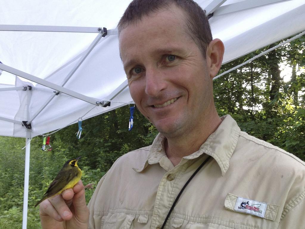 A male professor in a tan button up smiles at the camera while holding up a small, yellow and gray bird resting on his hand