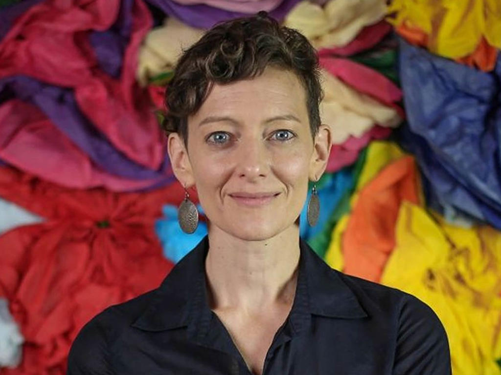 smiling professor in black blouse in front of a background made out of red, yellow, blue and orange cloth