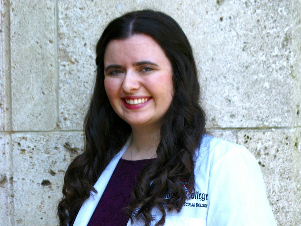 a headshot of a young white woman standing in front of a cement-tiled wall