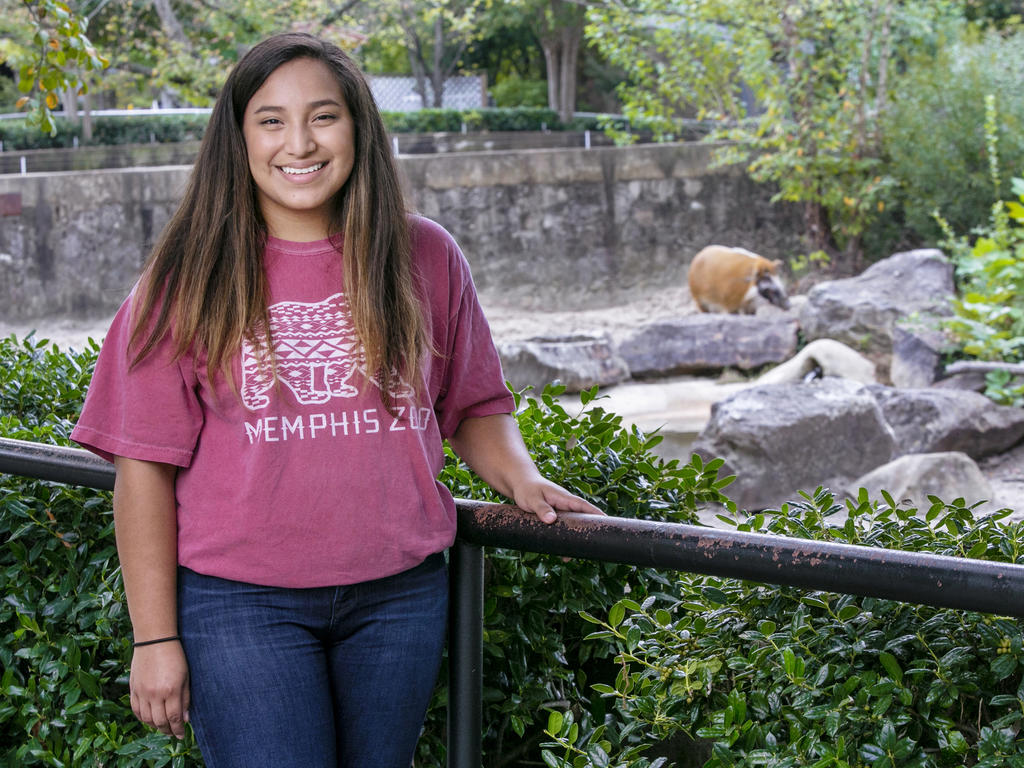 a young female student standing in front of a zoo exhibit in a t-shirt and jeans