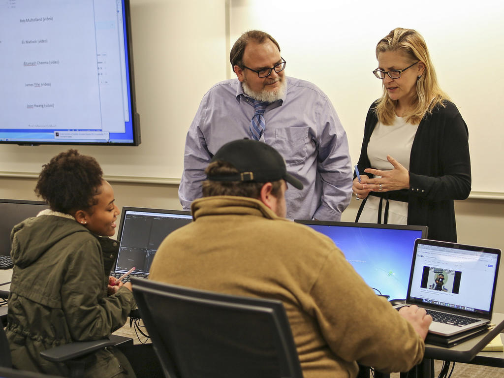 Two professors stand over a table at which two student sit in front of computers