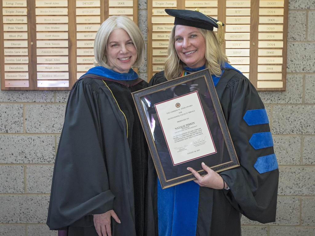 college administrator and professor dressed in black academic regalia and standing in front of a gold placard