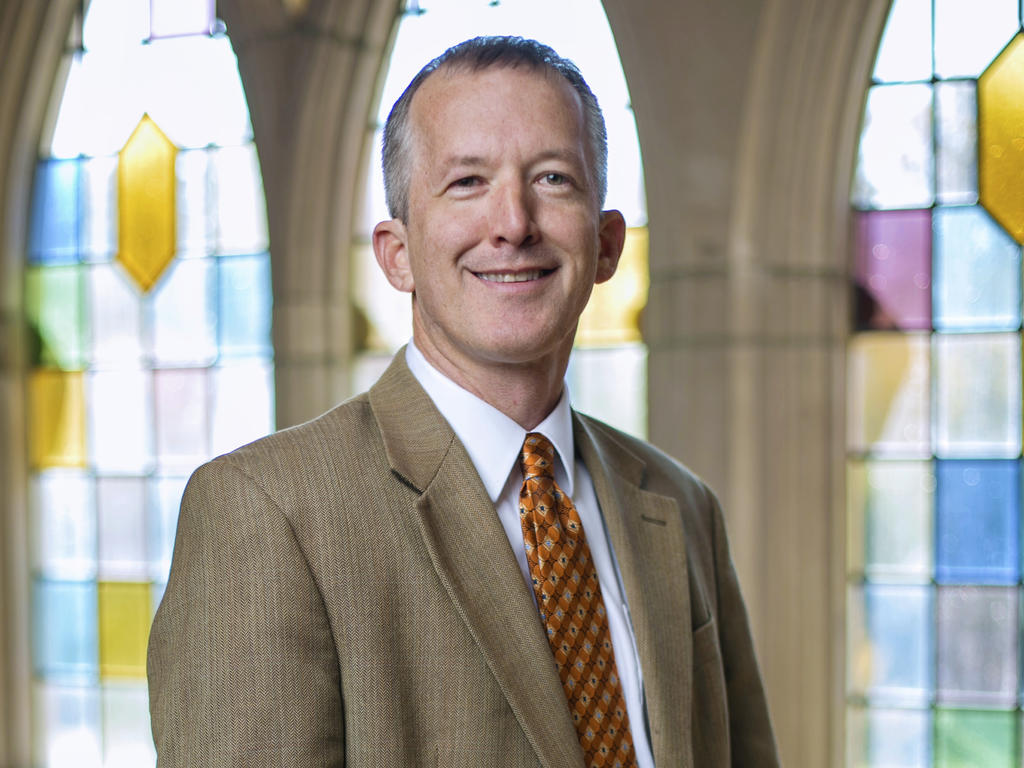 a middle-aged, white, male professor smiling in front of large, stained glass windows