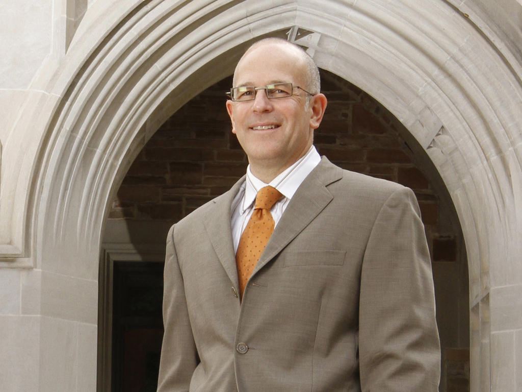 an older white male professor, who is bald and wearing a suit, standing in front of a gothic archway