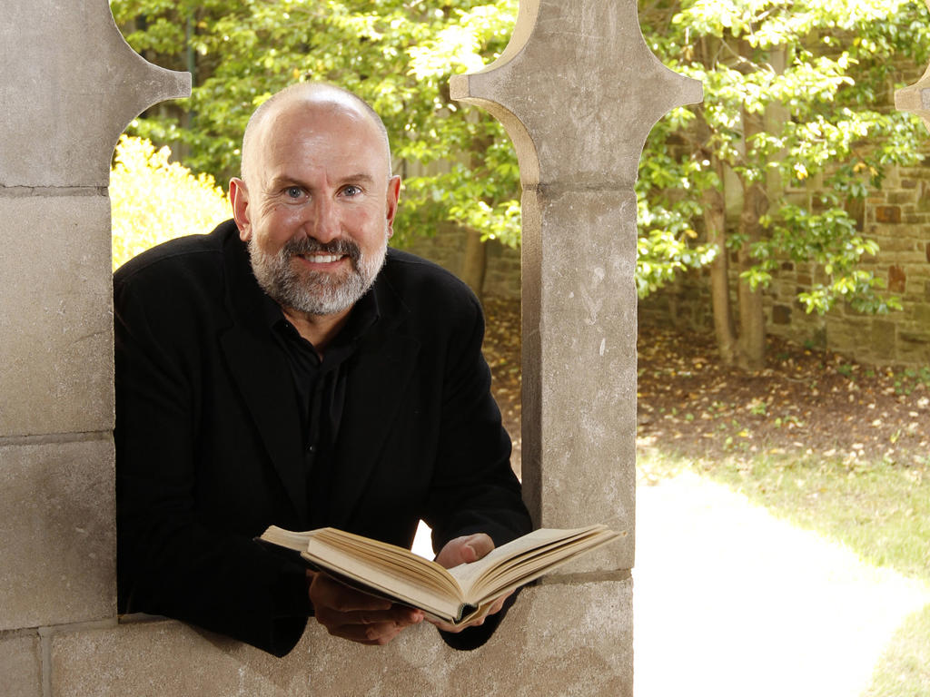 a middle-aged bald white man leaning out of a window while holding a book